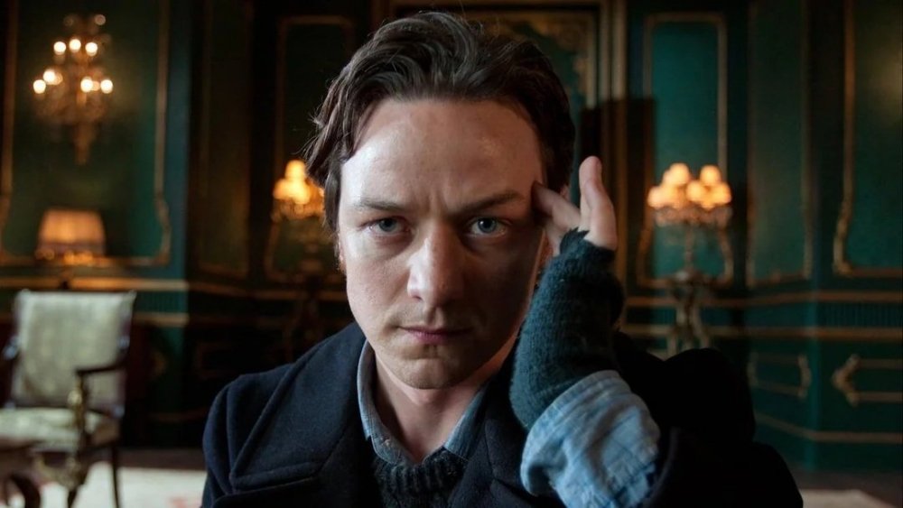 A man with tousled hair and a serious expression is seen in a richly decorated room, focusing intently with one hand on his forehead.